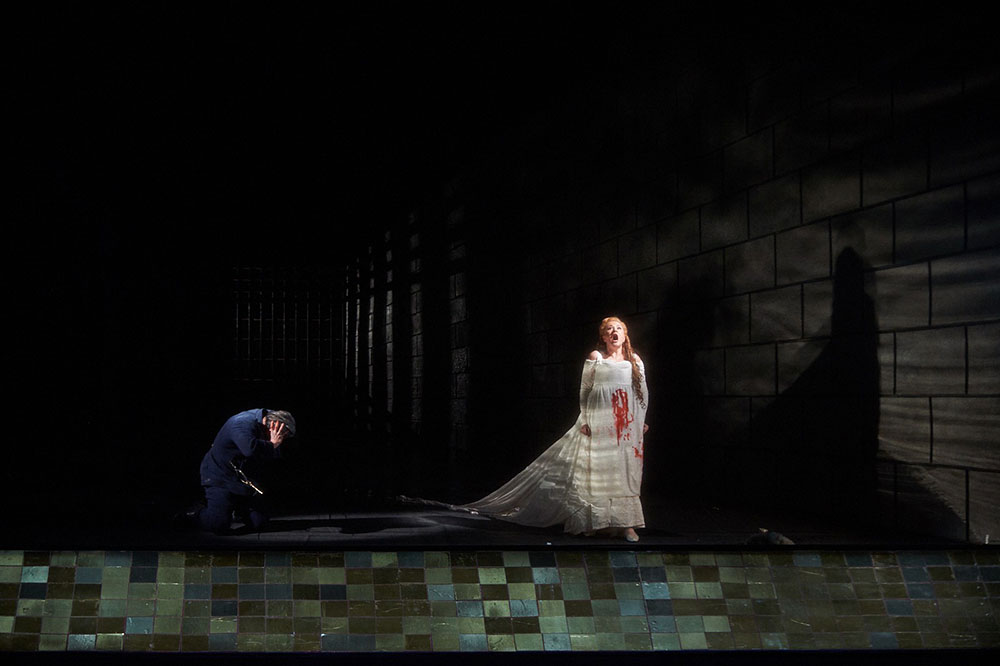 Production photo from Bluebeard's Castle, Canadian Opera Company, 2015, which shows a woman in a blood-stained white gown stands singing under a spotlight as a man kneels in shadow beside her in a dark, dramatic stage setting.