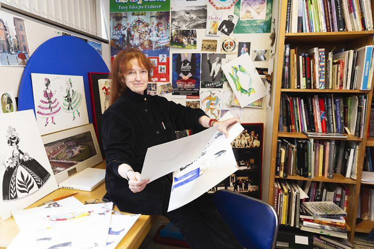 This image shows an artist in their studio, surrounded by colorful posters and bookshelves filled with various works. The artist, with red hair, is seated at a desk, holding a large piece of artwork in their hands. The space around them is filled with sketches and drawings, offering a glimpse into their creative process. The walls are decorated with a mix of promotional posters and illustrations, reflecting the vibrant, artistic environment.
