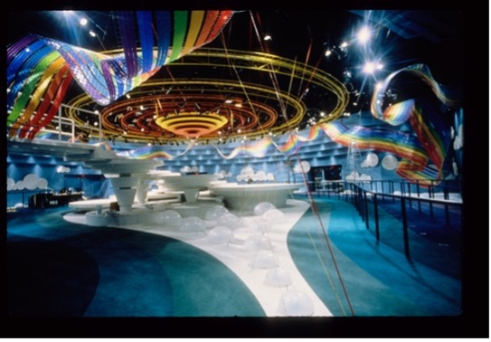 The photo captures an imaginative and colorful interior space. Above, swirling rainbow ribbons and concentric circles hang dramatically from the ceiling, while the floor features organic shapes, curves, and white structures resembling clouds. Bright, playful lighting complements the dreamy and whimsical mood of the environment.