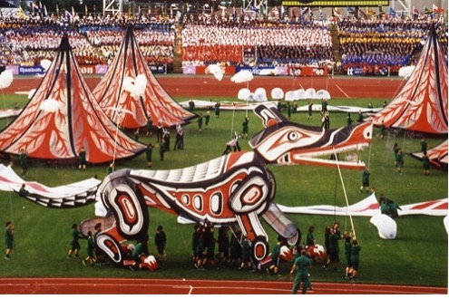 The image shows an outdoor performance scene featuring a large, stylized wolf puppet decorated in vibrant colors and tribal-inspired patterns. Several performers dressed in green hold and manipulate the puppet. Surrounding the wolf are large tent-like structures with decorative red and white designs, contributing to a lively and festive atmosphere.
