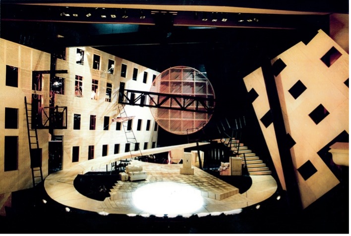 This image features an elaborate, architecturally striking theater set. It includes large, angled walls with multiple square windows, staircases, and ladders, giving it an industrial yet surreal appearance. At the center is a prominent circular structure with geometric patterns, crossed by a metal bridge-like structure. The design combines dramatic lighting and sharp angles to create a dynamic and visually engaging performance space.