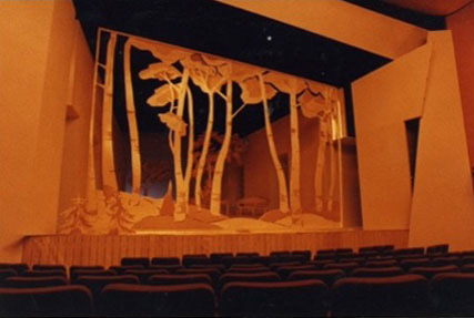 This image includes two distinct theatrical stage designs. The upper scene presents stylized cut-out trees and foliage, creating a forest-like environment. The lower design features tall, sculptural pine trees along an elegantly lit corridor, giving the stage depth and a tranquil, natural setting. Both scenes are characterized by warm, gentle lighting and simple yet expressive scenic elements, creating inviting, atmospheric settings within the theater space.