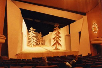 This image includes two distinct theatrical stage designs. The upper scene presents stylized cut-out trees and foliage, creating a forest-like environment. The lower design features tall, sculptural pine trees along an elegantly lit corridor, giving the stage depth and a tranquil, natural setting. Both scenes are characterized by warm, gentle lighting and simple yet expressive scenic elements, creating inviting, atmospheric settings within the theater space.