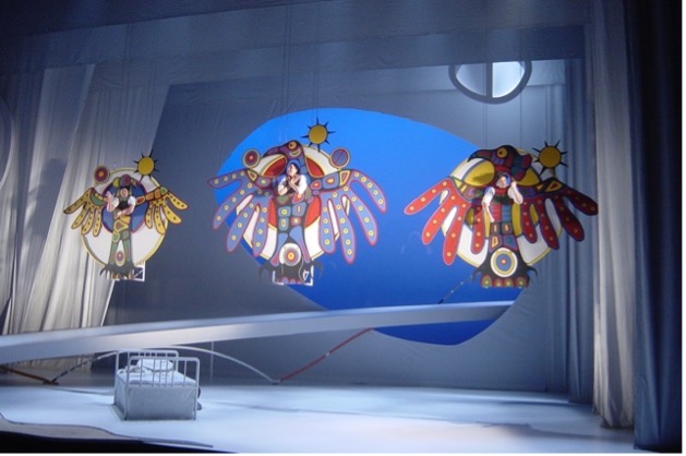 The image presents a theatrical stage with three performers suspended in stylized, colorful bird-like structures. Each performer is situated within intricately painted bird cutouts decorated with bright colors and symbolic imagery. The background is minimalist, featuring a curved platform, simple bed-like structure, and a vivid blue backdrop that enhances the overall surreal and imaginative quality of the scene.