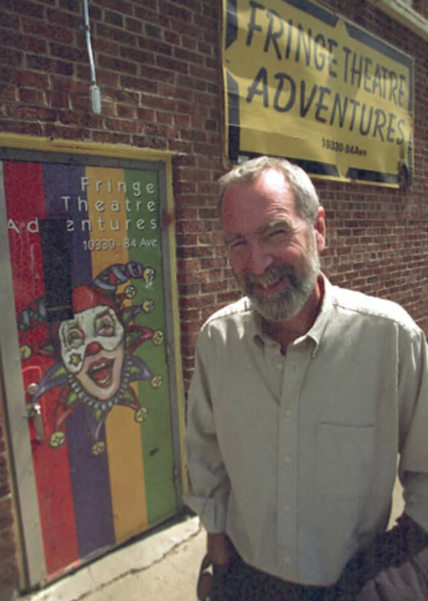 Brian Paisley, a smiling man with a gray beard and button-up shirt, standing outdoors in front of a red brick building featuring a painted door with a jester mask and a sign reading 'Fringe Theatre Adventures'.