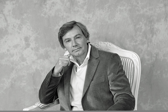 A black and white portrait of Don Shipley sitting in a white, ornate chair. He is wearing a dark corduroy blazer over a light-colored V-neck sweater and collared shirt.
