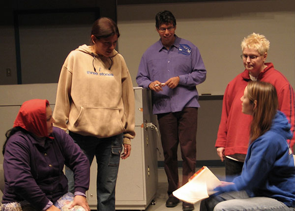 A group of five people is gathered indoors, seemingly in a casual or rehearsal setting. Charlene Hellson sits on the left, wearing a red headscarf and purple jacket, watching the interaction. Stacy da Silva, in a tan hoodie, stands in the center, looking down and engaging with a seated medical student in a blue hoodie who is holding a folder or papers. Dr. Lindsay Crowshoe stands in the background, observing or participating in the discussion, while Michele Decottignies, in a red hoodie with short blond hair, stands nearby. The mood appears focused and collaborative.